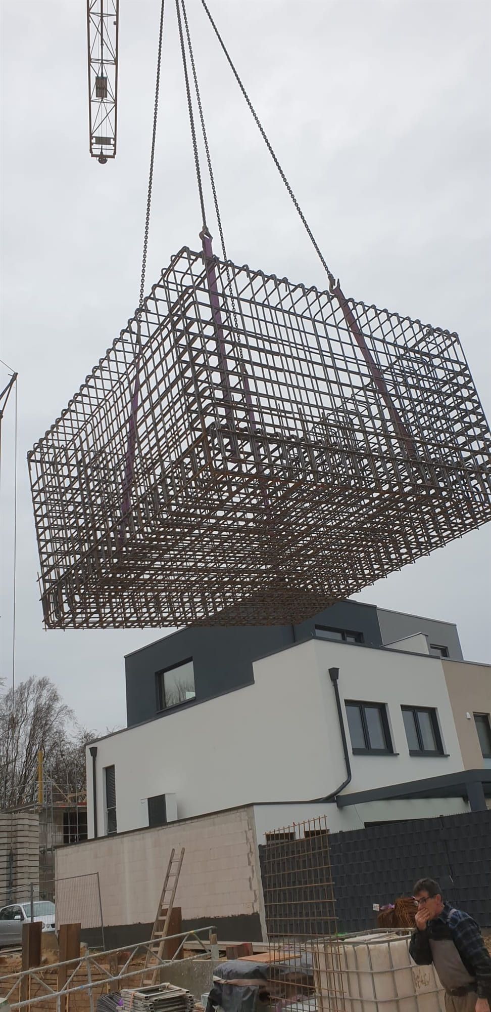 Ein großer Bewehrungskorb aus Betonstahl hängt am Kranhaken über einem Wohngebäude